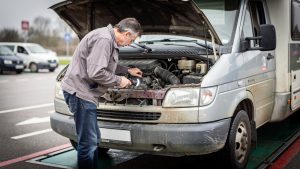 Un van aménagé d'occasion est stationné sur une aire de contrôle, le capot ouvert, tandis qu'un homme examine attentivement le moteur avec une lampe.